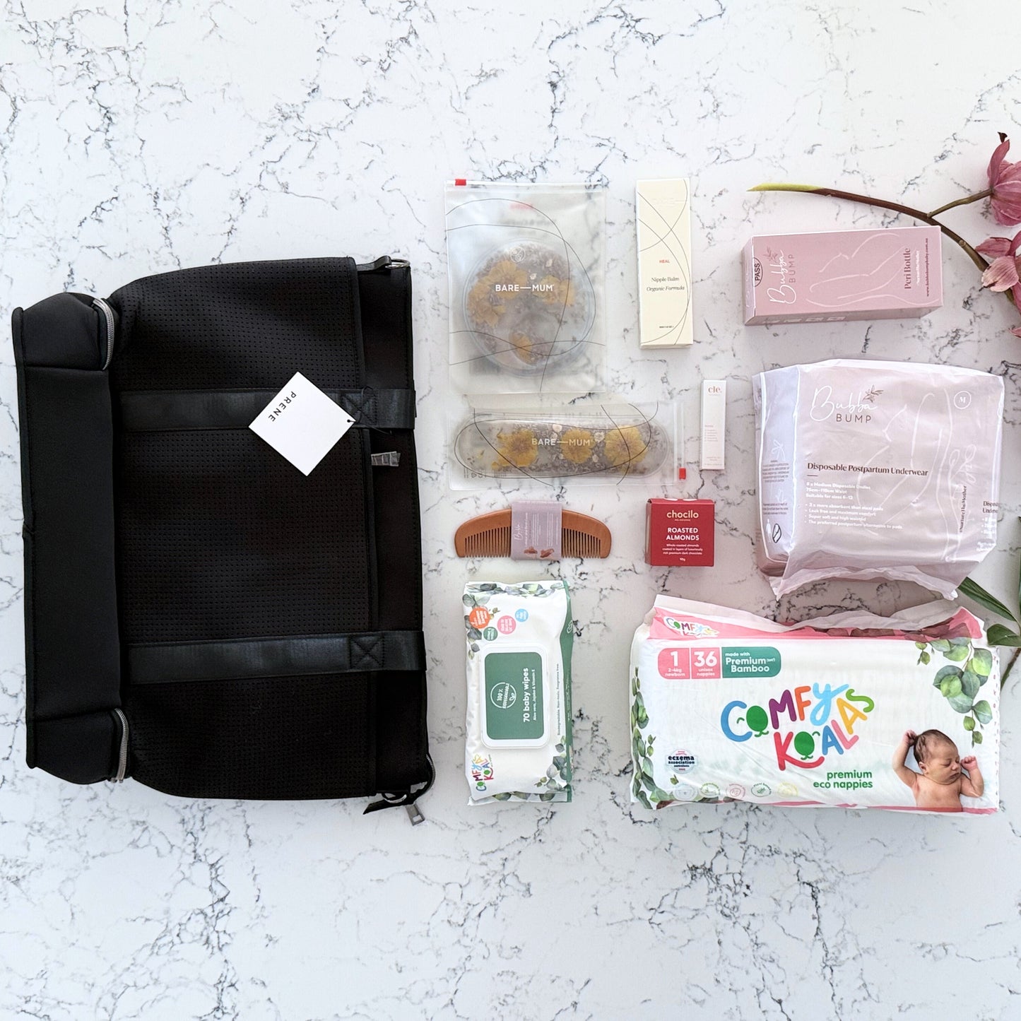 Omoiyari Hospital Bag with Prene Saturday Bag with bamboo nappies and wipes, perineal bottle, nipple cream, Essentials Oils roller, Postpartum disposable underwear, bra cooling inserts and chocolate on a marble surface with flowers.