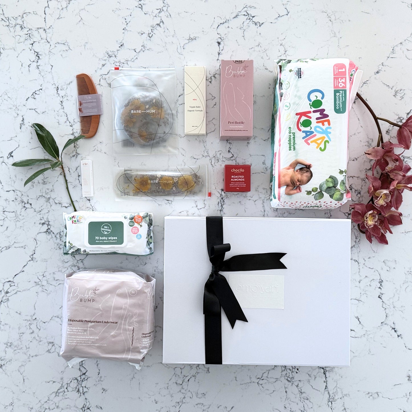 Omoiyari Hospital Bag gift hamper box  with bamboo nappies and wipes, perineal bottle, nipple cream, Essentials Oils roller, Postpartum disposable underwear, bra cooling inserts and chocolate on a marble surface with flowers.