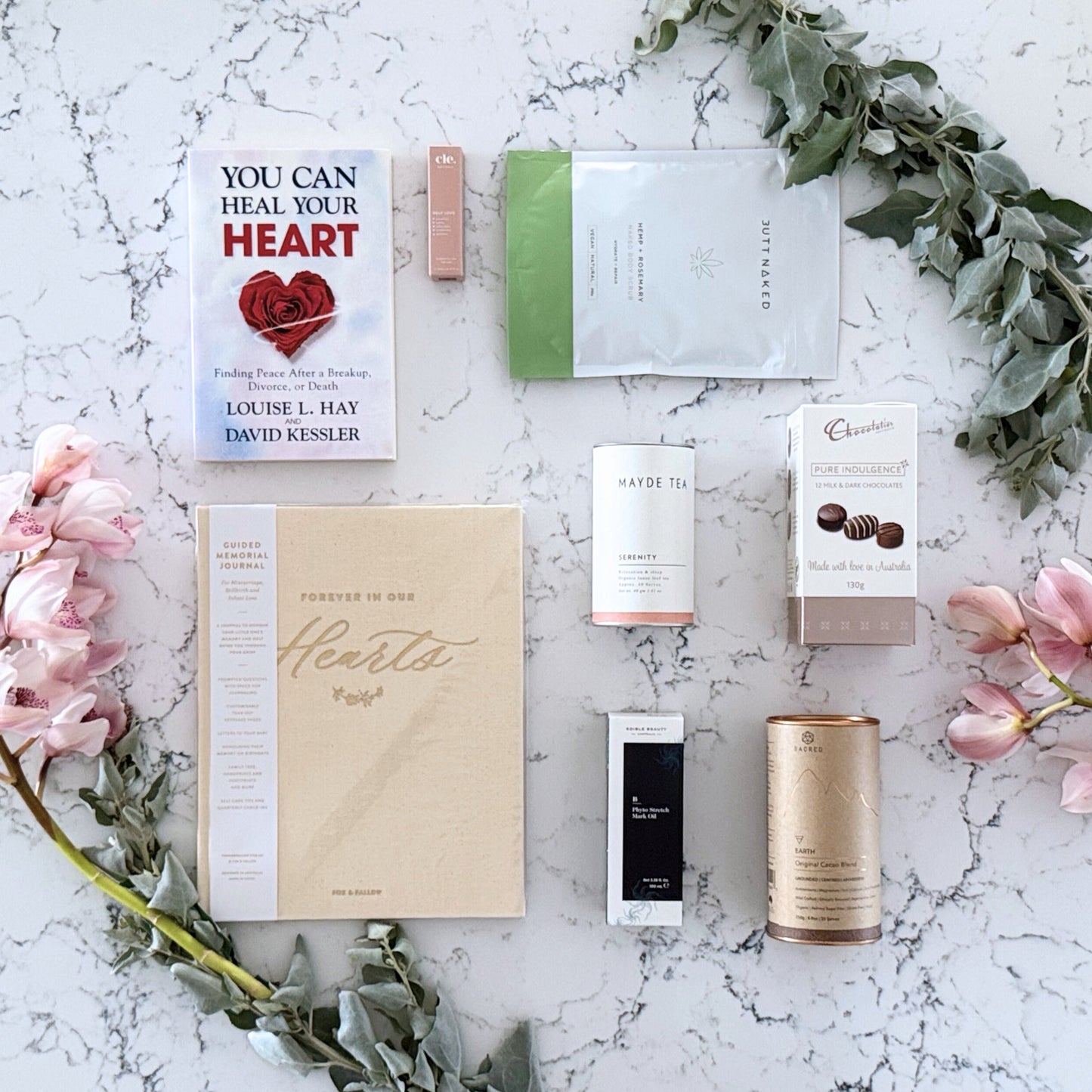 Omoiyari Healing Heart gift hamper box  with self-help book, In memory journal, Body scrub, organic tea, Essentials Oil, Chocolate, and soy candle on a marble surface with flowers.