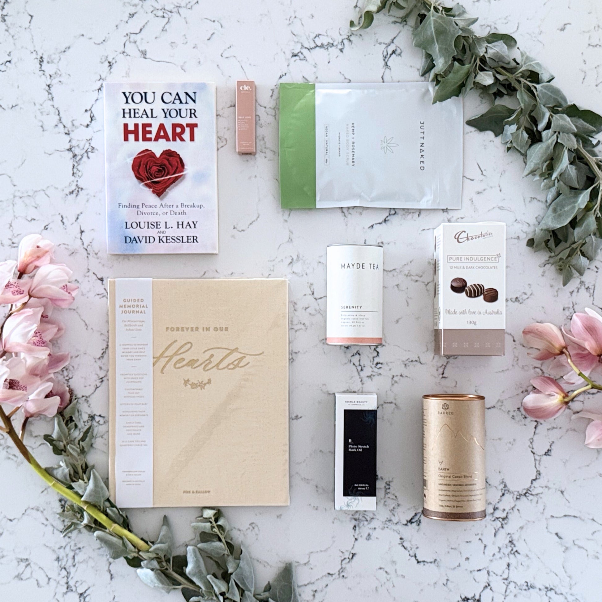 Omoiyari Healing Heart gift hamper box  with self-help book, In memory journal, Body scrub, organic tea, Essentials Oil, Chocolate, and soy candle on a marble surface with flowers.