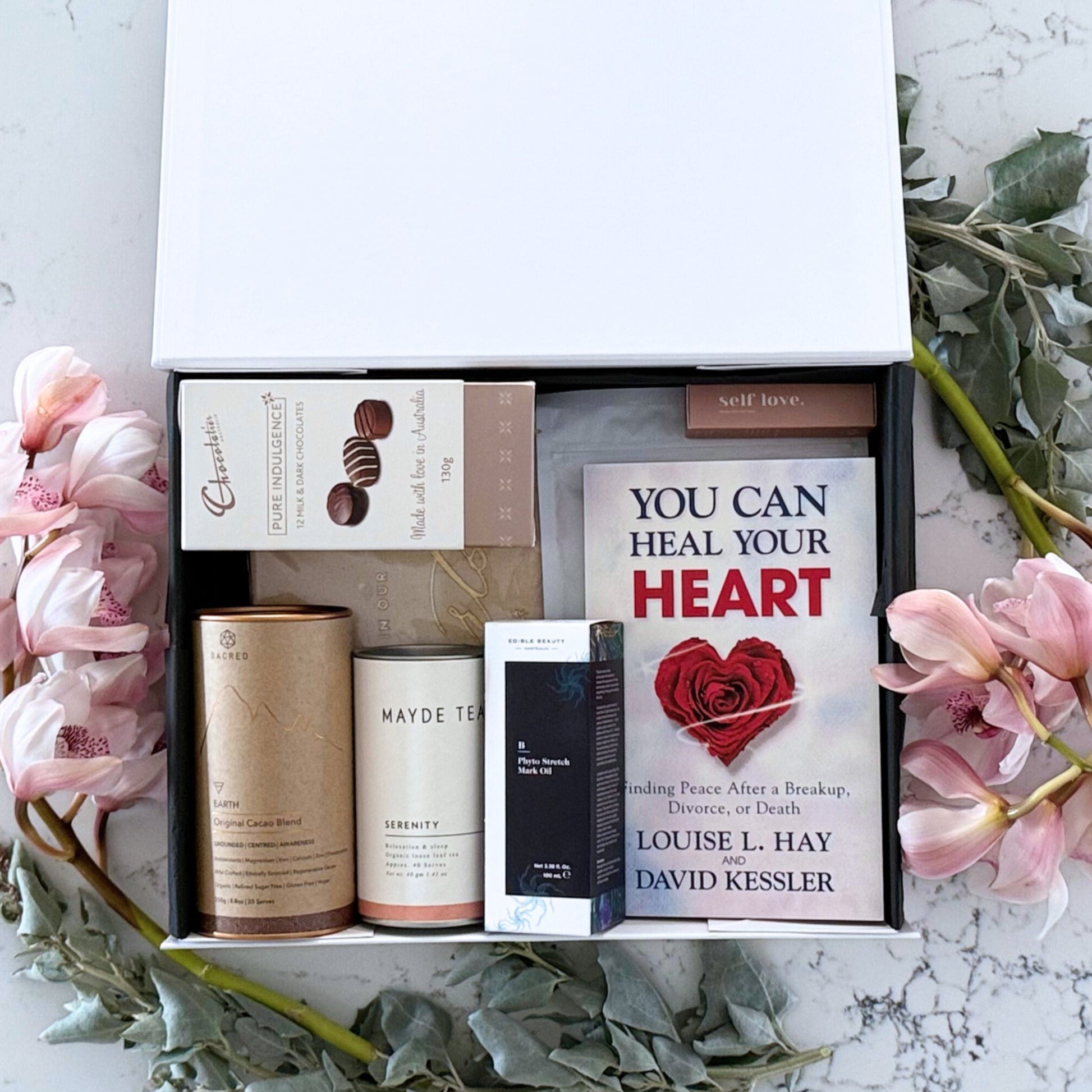Omoiyari Healing Heart gift hamper box  with self-help book, In memory journal, Body scrub, organic tea, Essentials Oil, Chocolate, and soy candle on a marble surface with flowers.