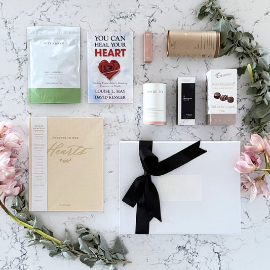 Omoiyari Healing Heart gift hamper box  with self-help book, In memory journal, Body scrub, organic tea, Essentials Oil, Chocolate, and soy candle on a marble surface with flowers.