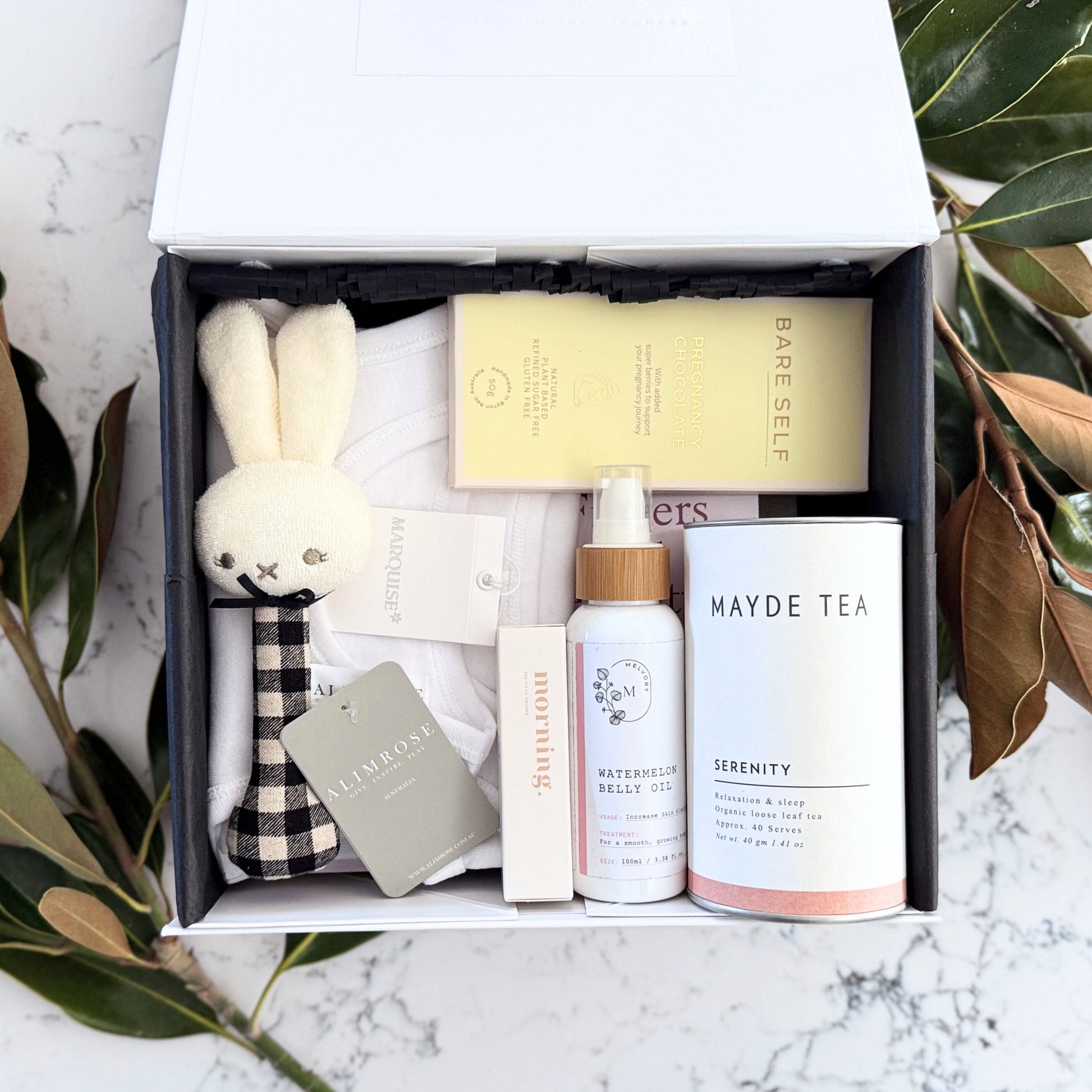 Omoiyari Mum & Baby gift hamper box - Skincare products, organic tea, stretch mark oil, essential oils, pregnancy journal, pregnancy chocolate and green magnolia leaves on a marble surface