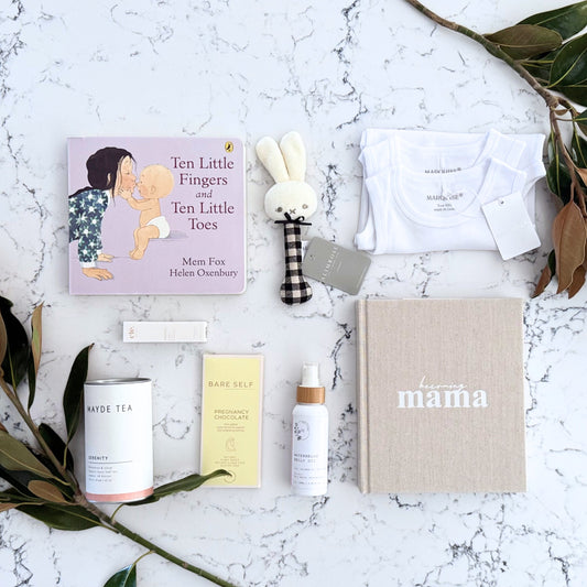 Omoiyari Mum & Baby gift hamper box - Skincare products, organic tea, stretch mark oil, essential oils, pregnancy journal, pregnancy chocolate and green magnolia leaves on a marble surface