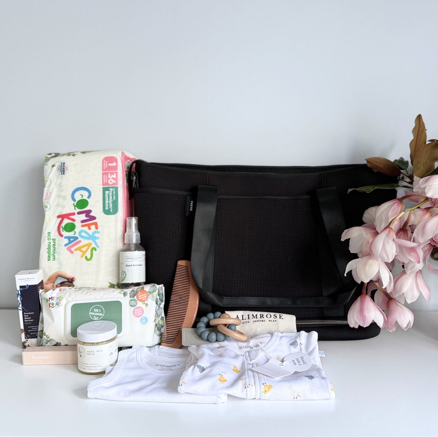 Omoiyari Nappy Bag Luxe with PRene Saturday Bag. Marquise babywear, bamboo nappies and wipes, bamboo comb, Hand cream, Teether ring, baby balm and sanitiser on a marble surface with magnolia leaves.