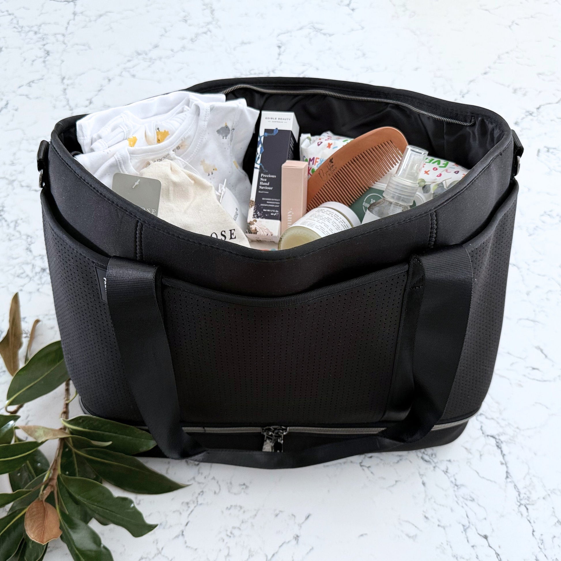 Omoiyari Nappy Bag Luxe with PRene Saturday Bag filled with Marquise babywear, bamboo nappies and wipes, bamboo comb, Hand cream, Teether ring, baby balm and sanitiser on a marble surface with magnolia leaves.