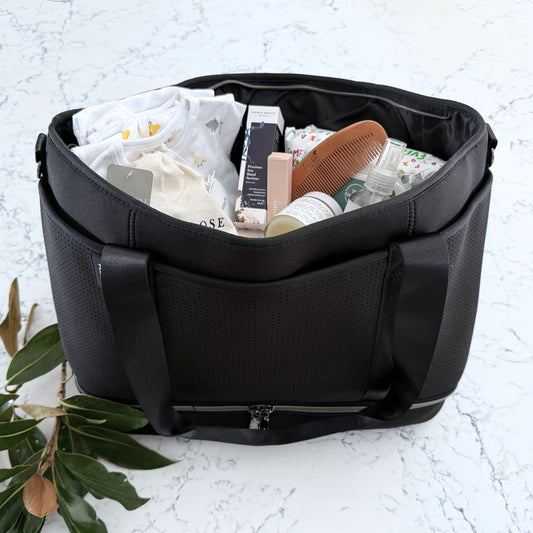 Omoiyari Nappy Bag Luxe with PRene Saturday Bag filled with Marquise babywear, bamboo nappies and wipes, bamboo comb, Hand cream, Teether ring, baby balm and sanitiser on a marble surface with magnolia leaves.