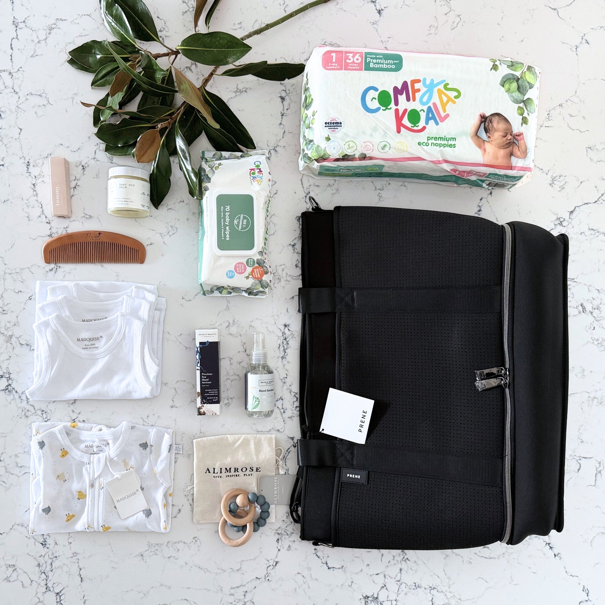 Omoiyari Nappy Bag Luxe with PRene Saturday Bag. Marquise babywear, bamboo nappies and wipes, bamboo comb, Hand cream, Teether ring, baby balm and sanitiser on a marble surface with magnolia leaves.