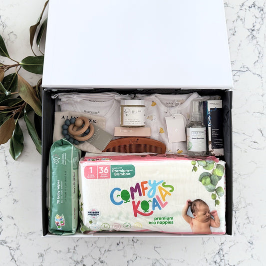 Omoiyari Nappy Bag Luxe Gift Hamper Box with Marquise babywear, bamboo nappies and wipes, bamboo comb, Hand cream, Teether ring, baby balm and sanitiser on a marble surface with magnolia leaves.