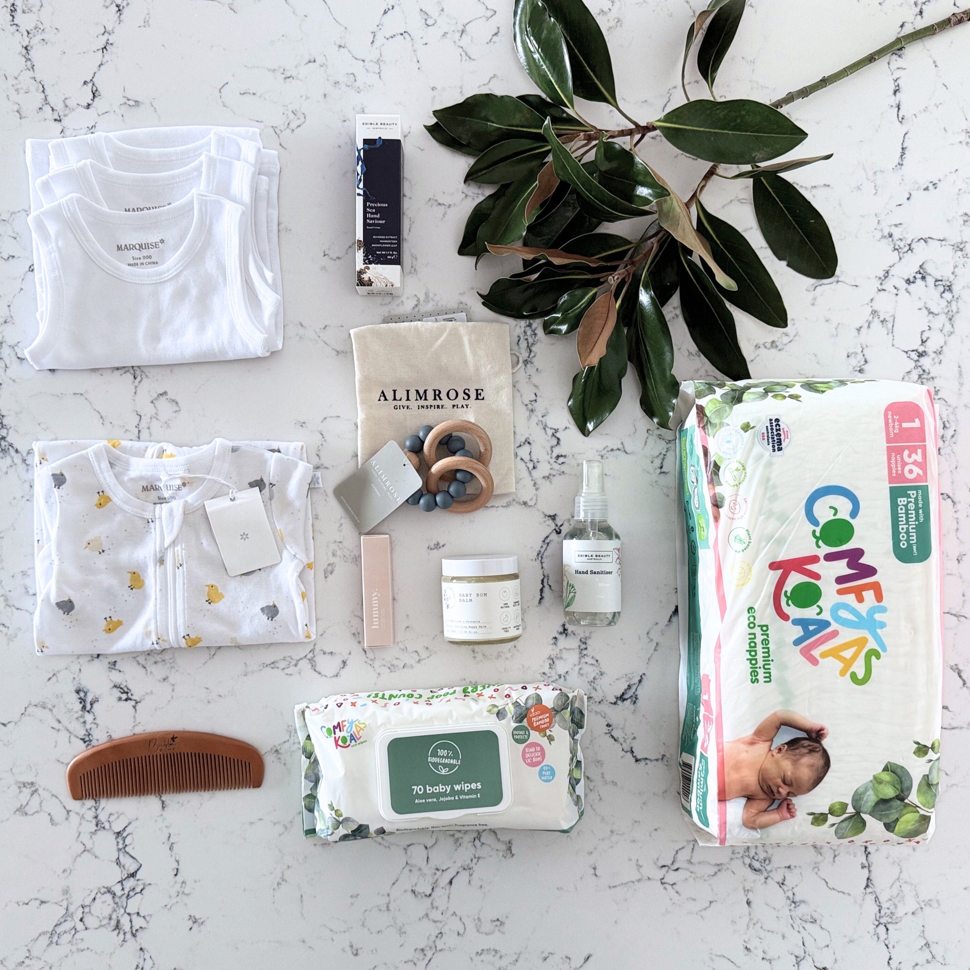 Omoiyari Nappy Bag Luxe Gift Hamper Box with Marquise babywear, bamboo nappies and wipes, bamboo comb, Hand cream, Teether ring, baby balm and sanitiser on a marble surface with magnolia leaves.