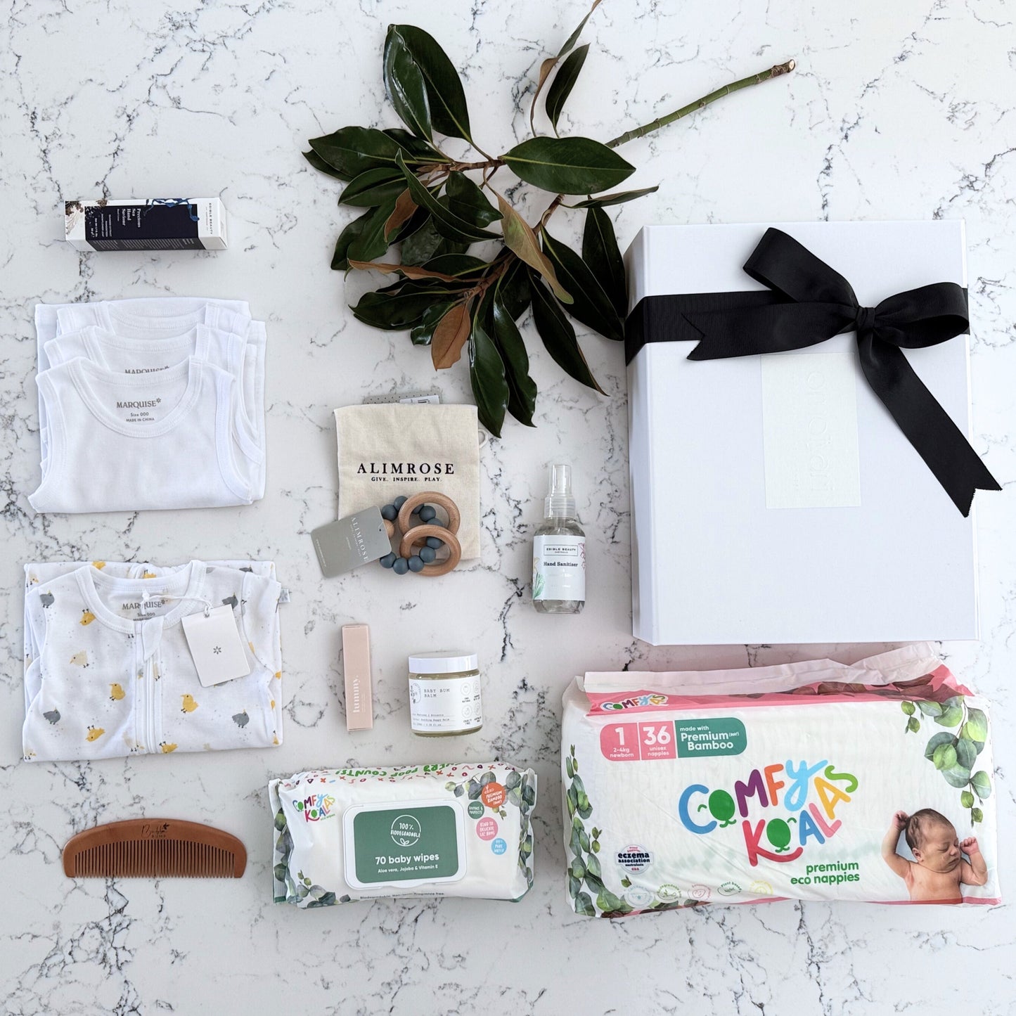 Omoiyari Nappy Bag Luxe Gift Hamper Box with Marquise babywear, bamboo nappies and wipes, bamboo comb, Hand cream, Teether ring, baby balm and sanitiser on a marble surface with magnolia leaves.