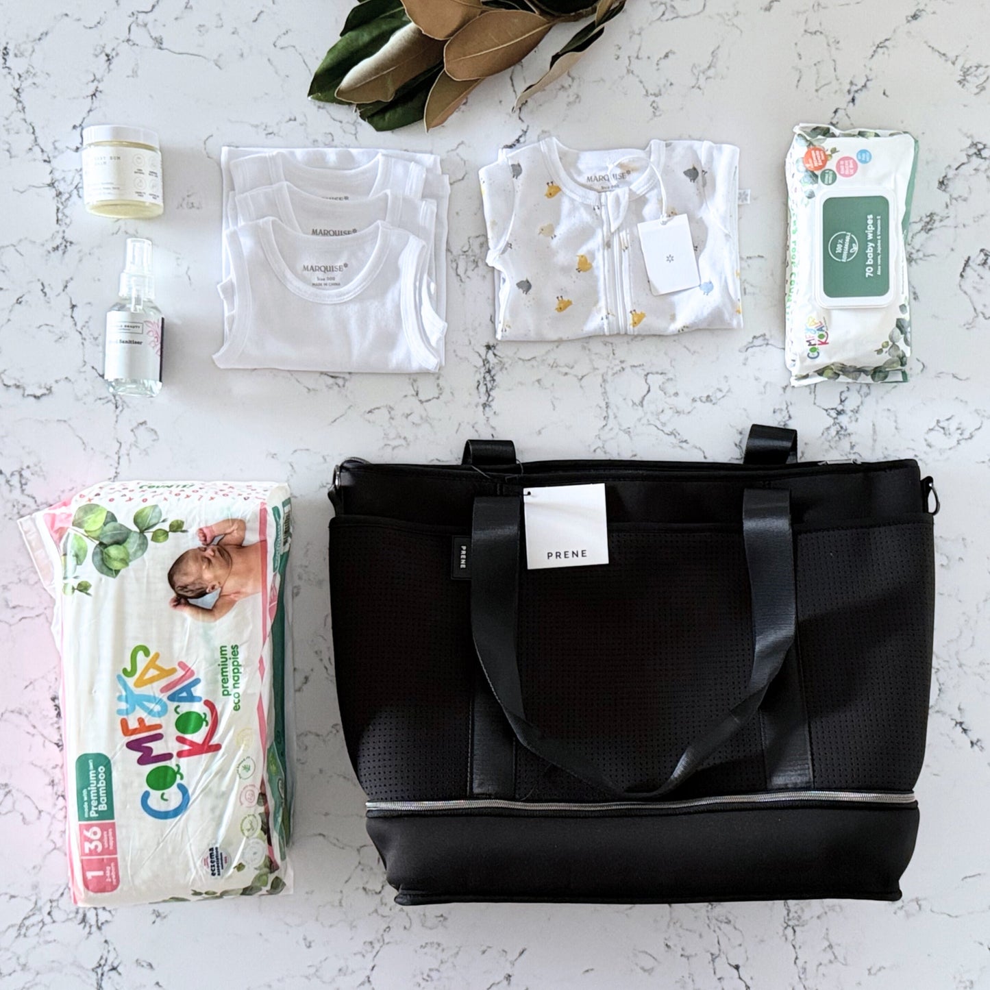 Omoiyari Nappy Bag with Prene Saturday Bag with Marquise babywear, bamboo nappies and wipes, baby balm and sanitiser on a marble surface with magnolia leaves.