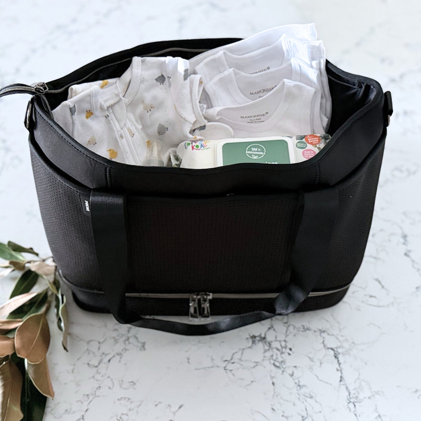 Omoiyari Nappy Bag with Prene Saturday Bag with Marquise babywear, bamboo nappies and wipes, baby balm and sanitiser on a marble surface with magnolia leaves.