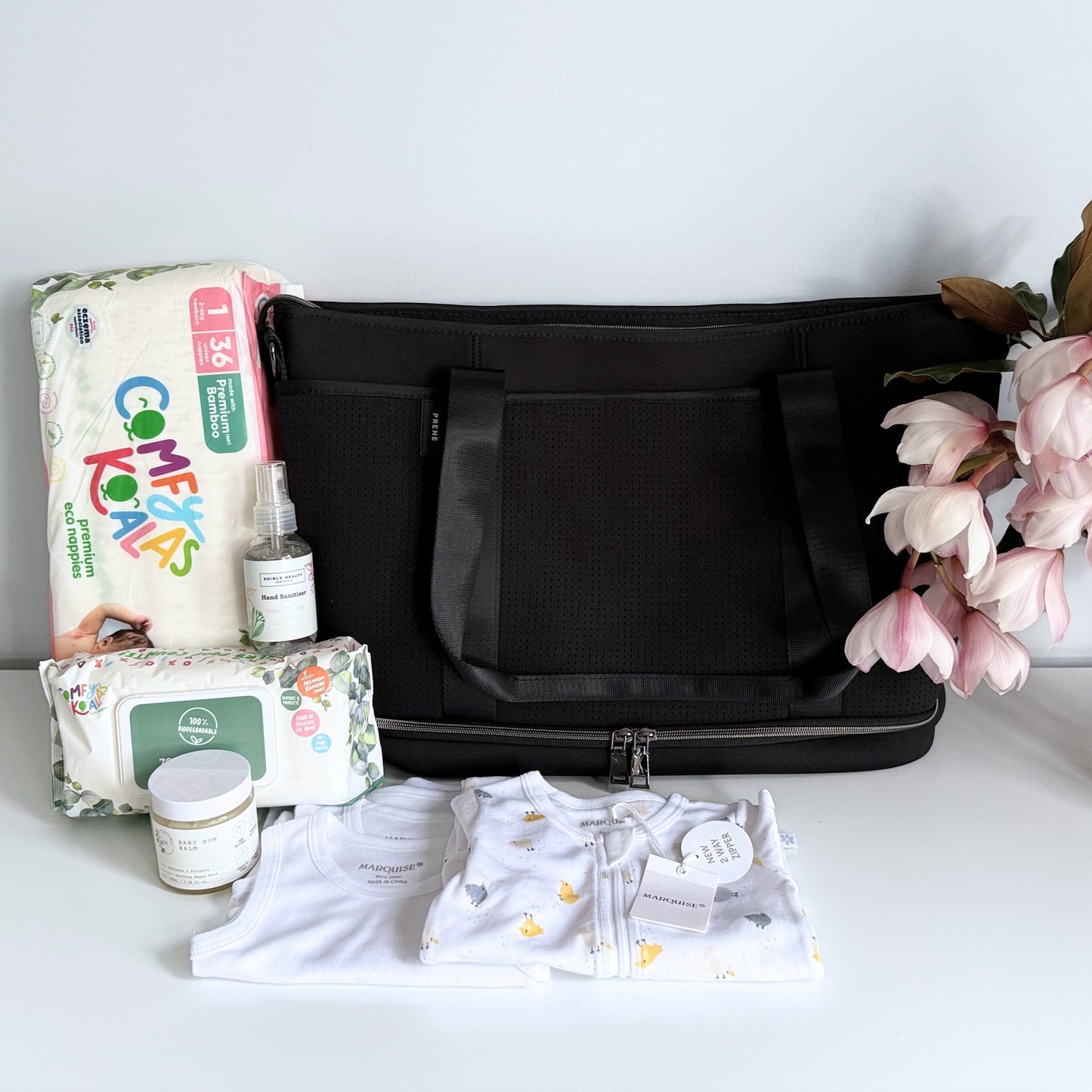 Omoiyari Nappy Bag with Prene Saturday Bag with Marquise babywear, bamboo nappies and wipes, baby balm and sanitiser on a marble surface with magnolia leaves.