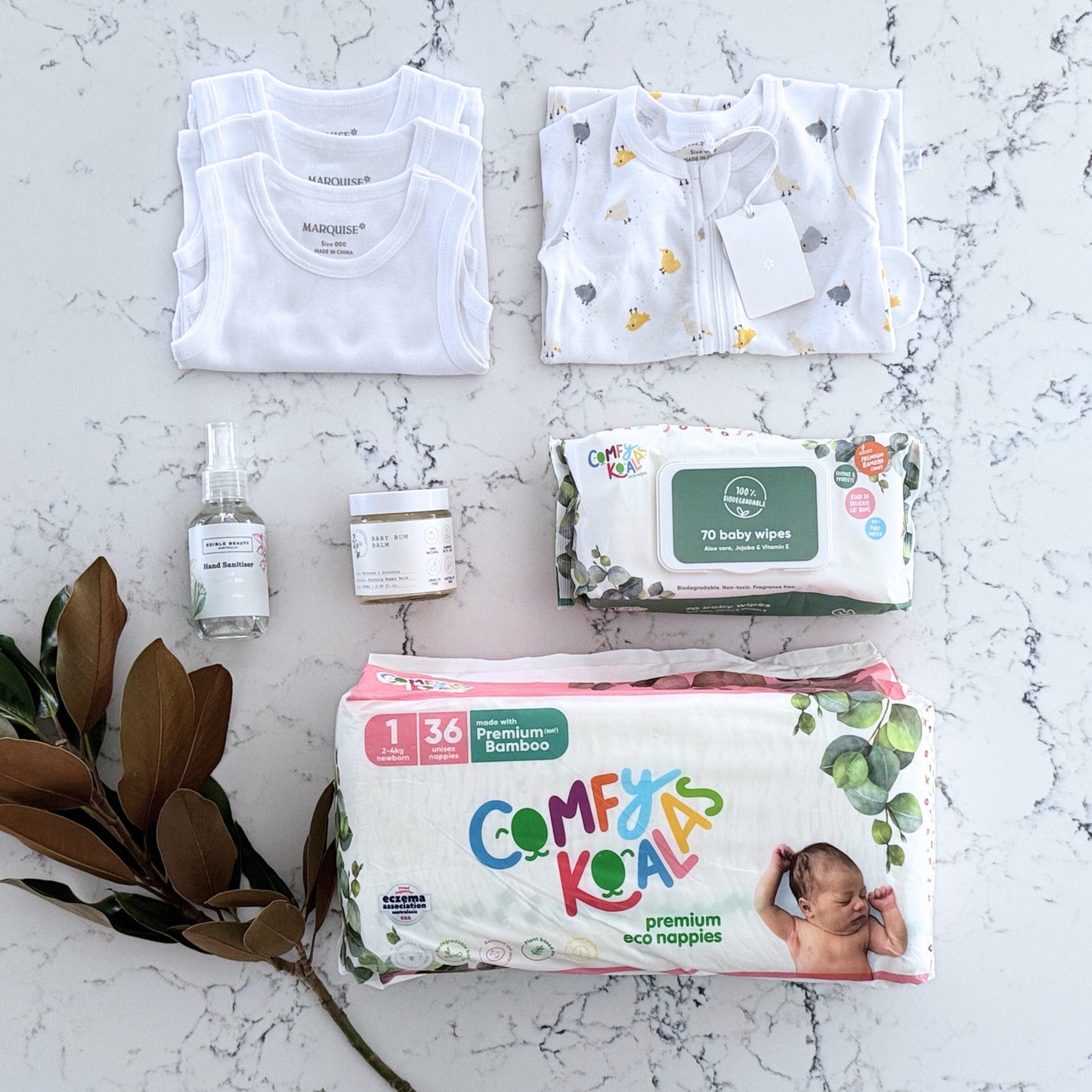 Omoiyari Nappy Bag Gift Hamper Box with Marquise babywear, bamboo nappies and wipes, baby balm and sanitiser on a marble surface with magnolia leaves.