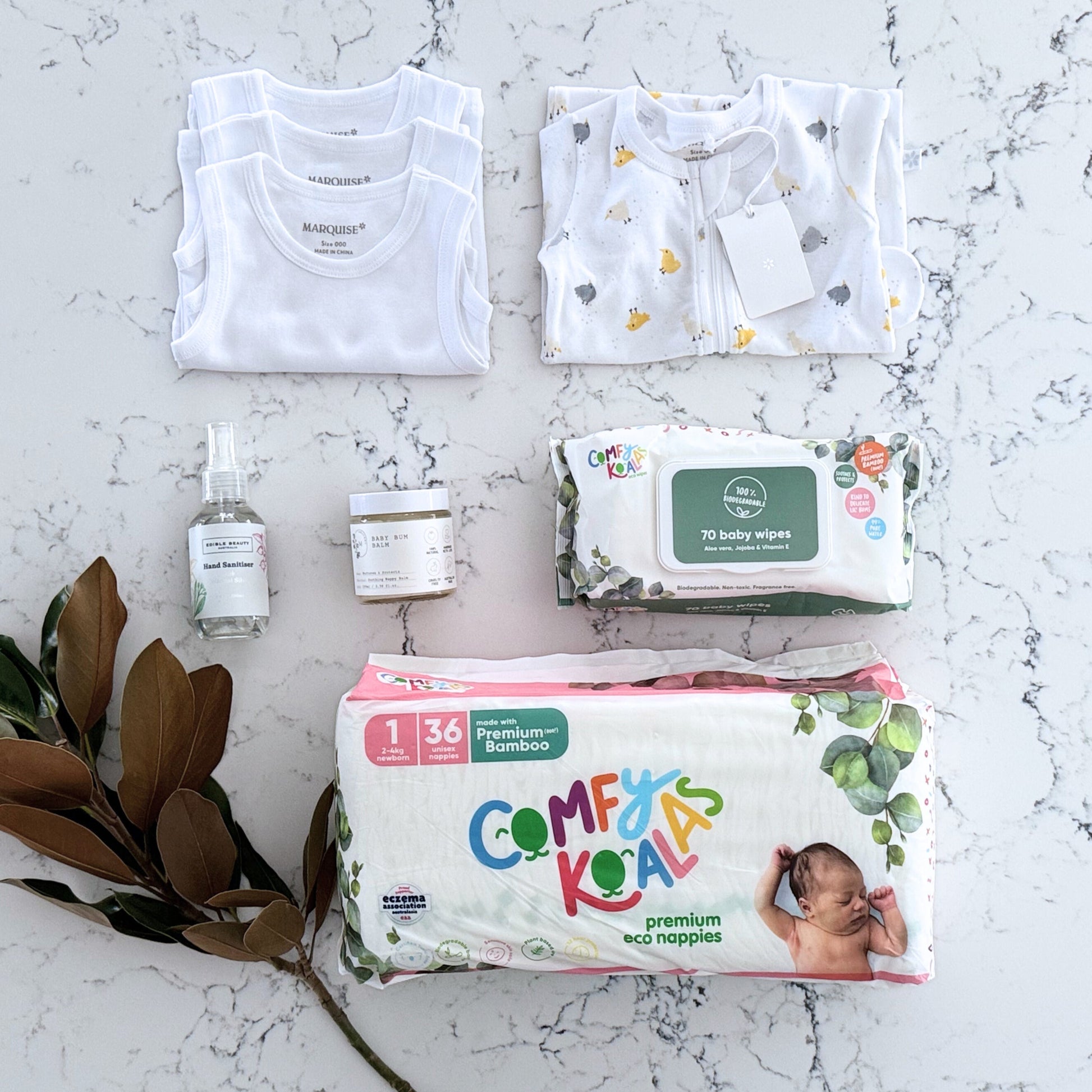 Omoiyari Nappy Bag Gift Hamper Box with Marquise babywear, bamboo nappies and wipes, baby balm and sanitiser on a marble surface with magnolia leaves.