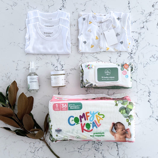 Omoiyari Nappy Bag Gift Hamper Box with Marquise babywear, bamboo nappies and wipes, baby balm and sanitiser on a marble surface with magnolia leaves.