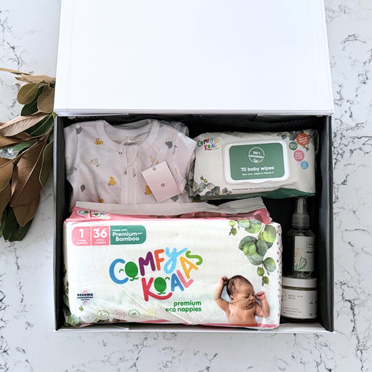 Omoiyari Nappy Bag Gift Hamper Box with Marquise babywear, bamboo nappies and wipes, baby balm and sanitiser on a marble surface with magnolia leaves.