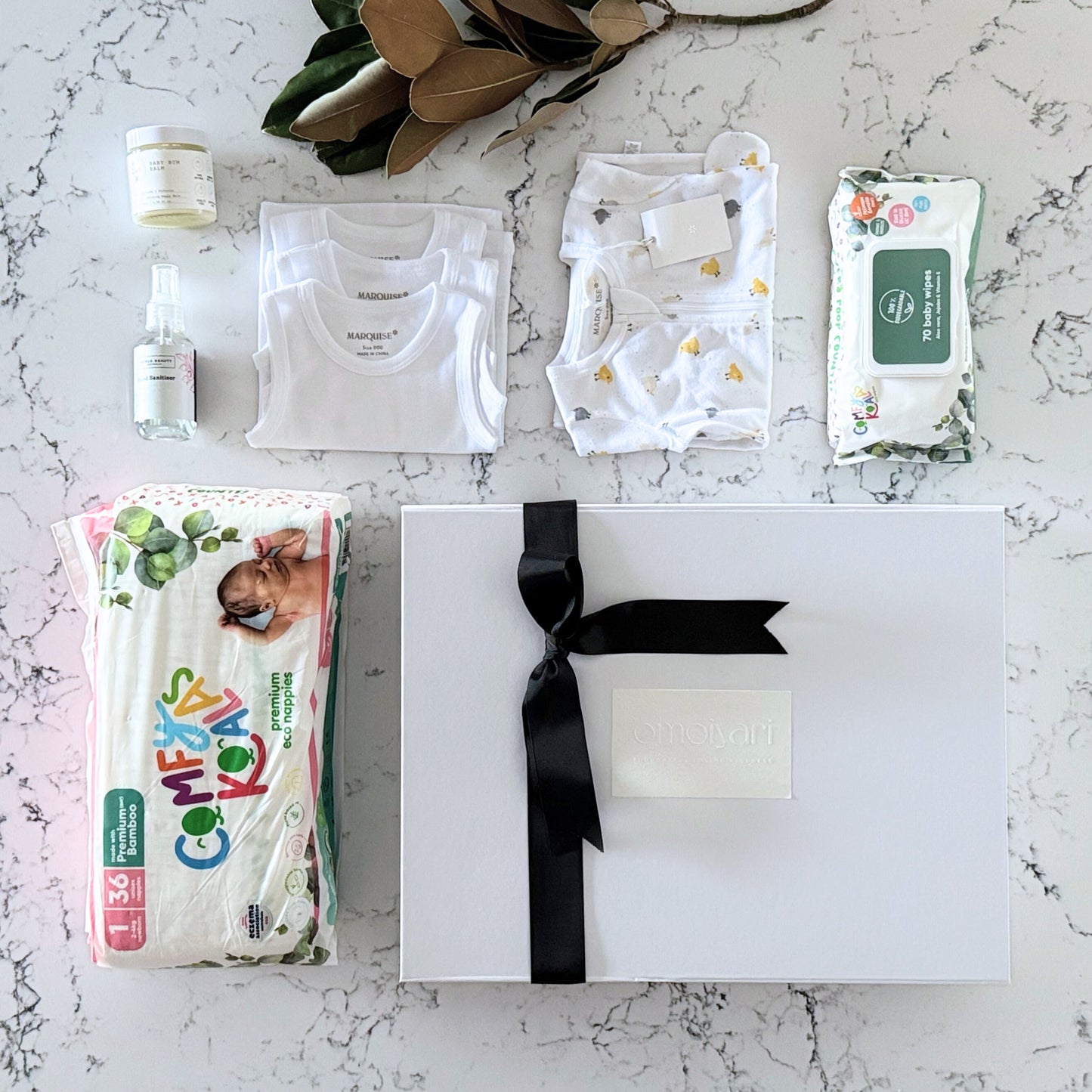 Omoiyari Nappy Bag Gift Hamper Box with Marquise babywear, bamboo nappies and wipes, baby balm and sanitiser on a marble surface with magnolia leaves.