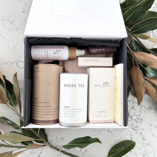 Omoiyari Nurture & Nourish Luxe gift hamper box - Skincare products, organic cacao and tea, pregnancy chocolate, stretch mark oil, radiance body oil, lip balm and green magnolia leaves on a marble surface