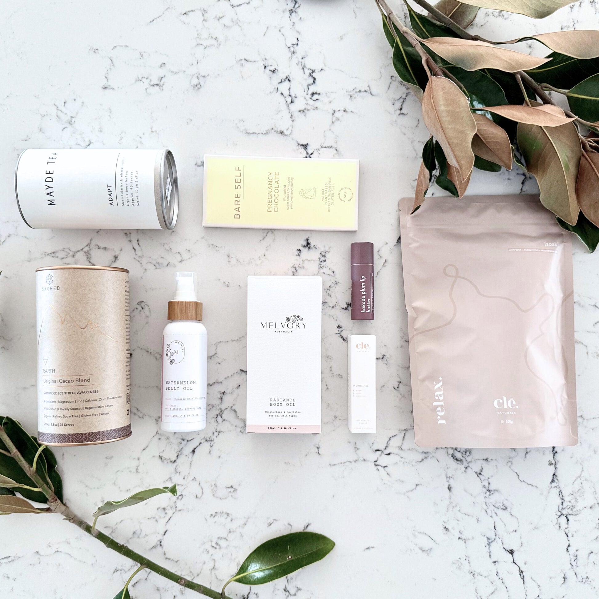 Omoiyari Nurture & Nourish Luxe gift hamper box - Skincare products, organic cacao and tea, pregnancy chocolate, stretch mark oil, radiance body oil, lip balm and green magnolia leaves on a marble surface