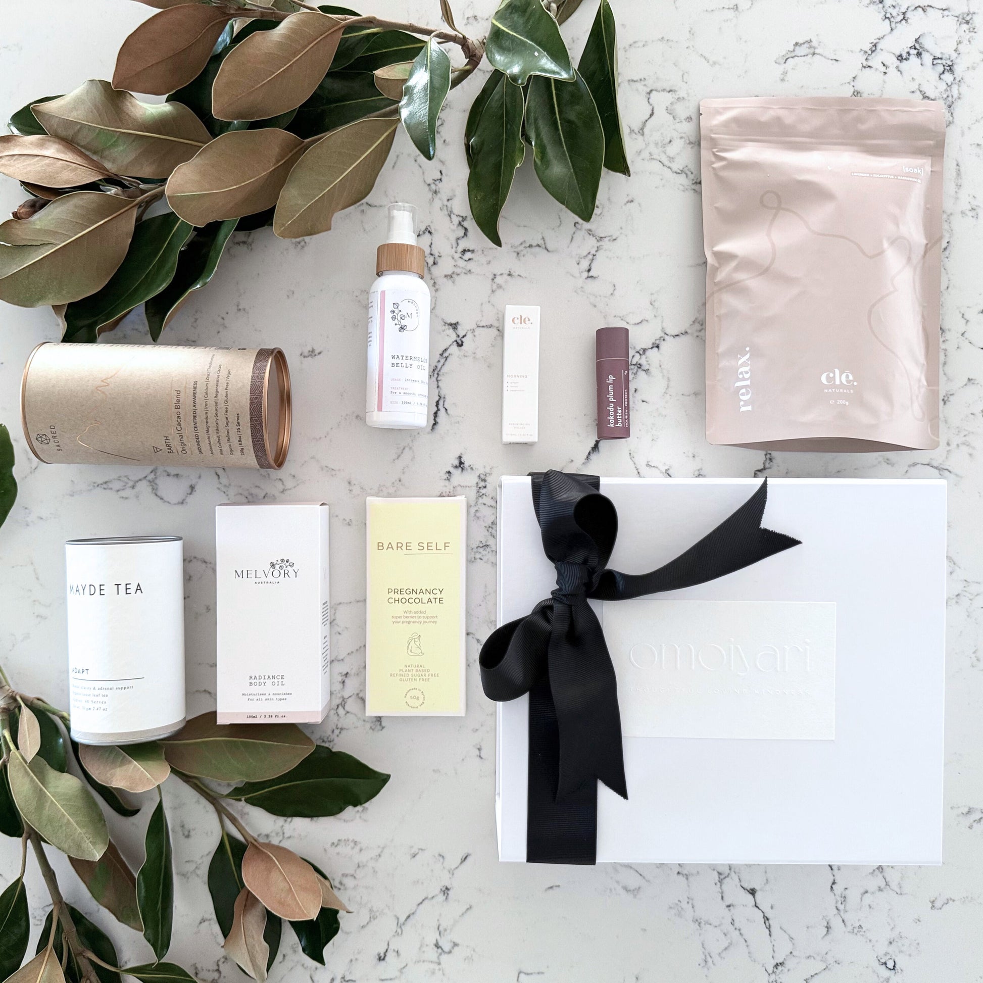 Omoiyari Nurture & Nourish Luxe gift hamper box - Skincare products, organic cacao and tea, pregnancy chocolate, stretch mark oil, radiance body oil, lip balm and green magnolia leaves on a marble surface