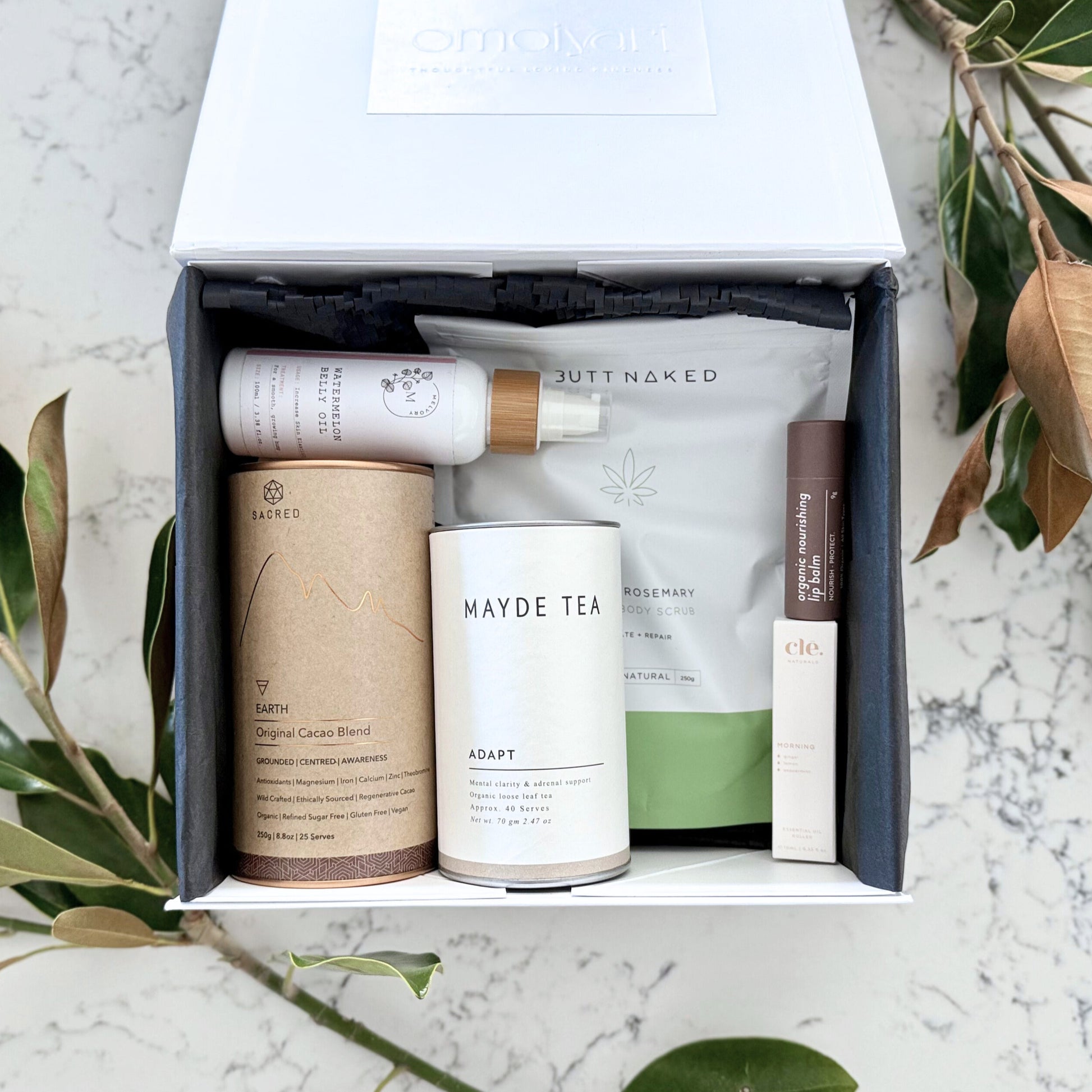 Omoiyari Nurture & Nourish gift hamper box - Skincare products, organic cacao and tea, stretch mark oil, lip balm and green magnolia leaves on a marble surface
