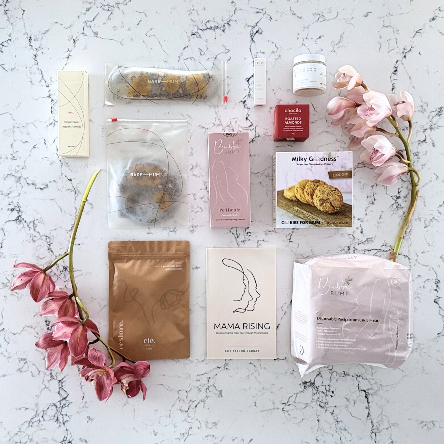 Omoiyari Postpartum Care gift hamper box - Perineal bottle, Sitz bath soak, postpartum underwear, Perineal insert, Lactation cookies, Roasted almond chocolate, nipple balm, Mama Rising book, Essential Oils roller, with pink orchid flowers on a marble surface