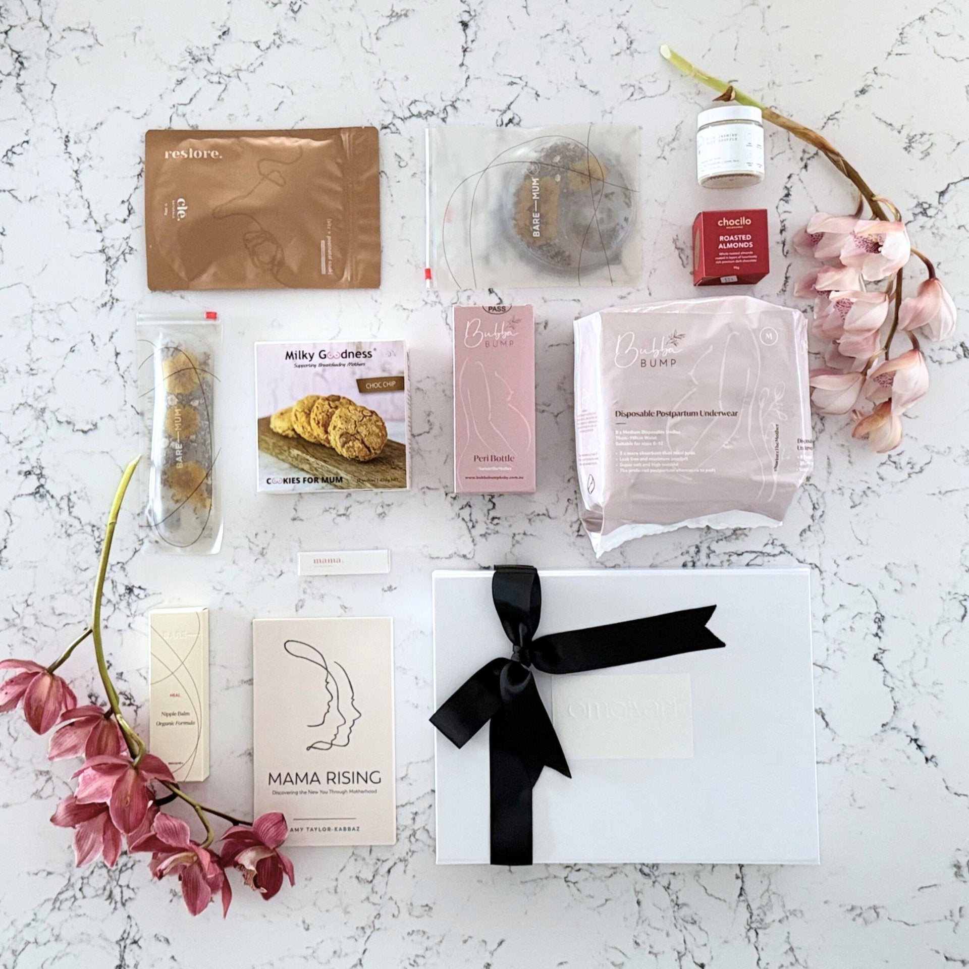 Omoiyari Postpartum Care gift hamper box - Perineal bottle, Sitz bath soak, postpartum underwear, Perineal insert, Lactation cookies, Roasted almond chocolate, nipple balm, Mama Rising book, Essential Oils roller, with pink orchid flowers on a marble surface