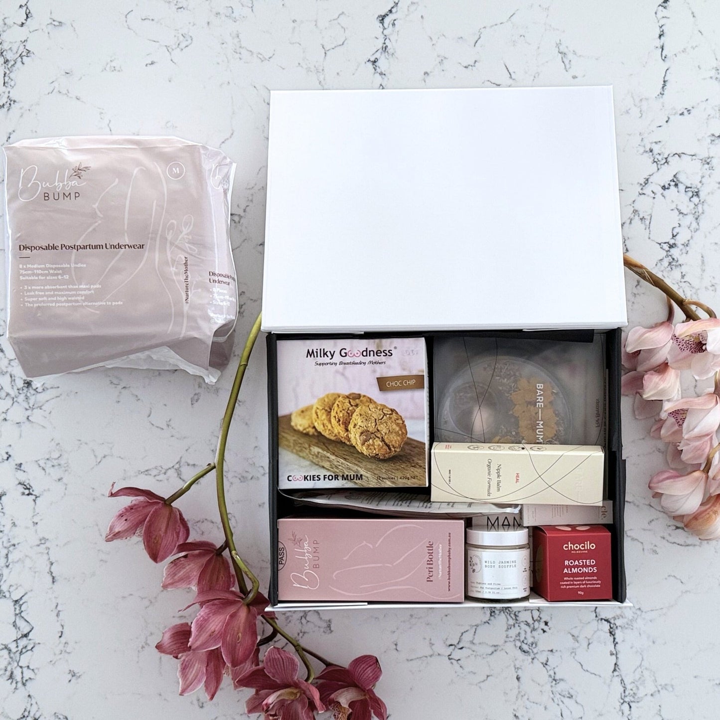Omoiyari Postpartum Care gift hamper box - Perineal bottle, Sitz bath soak, postpartum underwear, Perineal insert, Lactation cookies, Roasted almond chocolate, nipple balm, Mama Rising book, Essential Oils roller, with pink orchid flowers on a marble surface