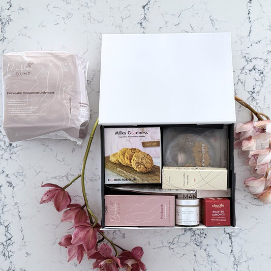 Omoiyari Postpartum Care gift hamper box - Perineal bottle, Sitz bath soak, postpartum underwear, Perineal insert, Lactation cookies, Roasted almond chocolate, nipple balm, Mama Rising book, Essential Oils roller, with pink orchid flowers on a marble surface