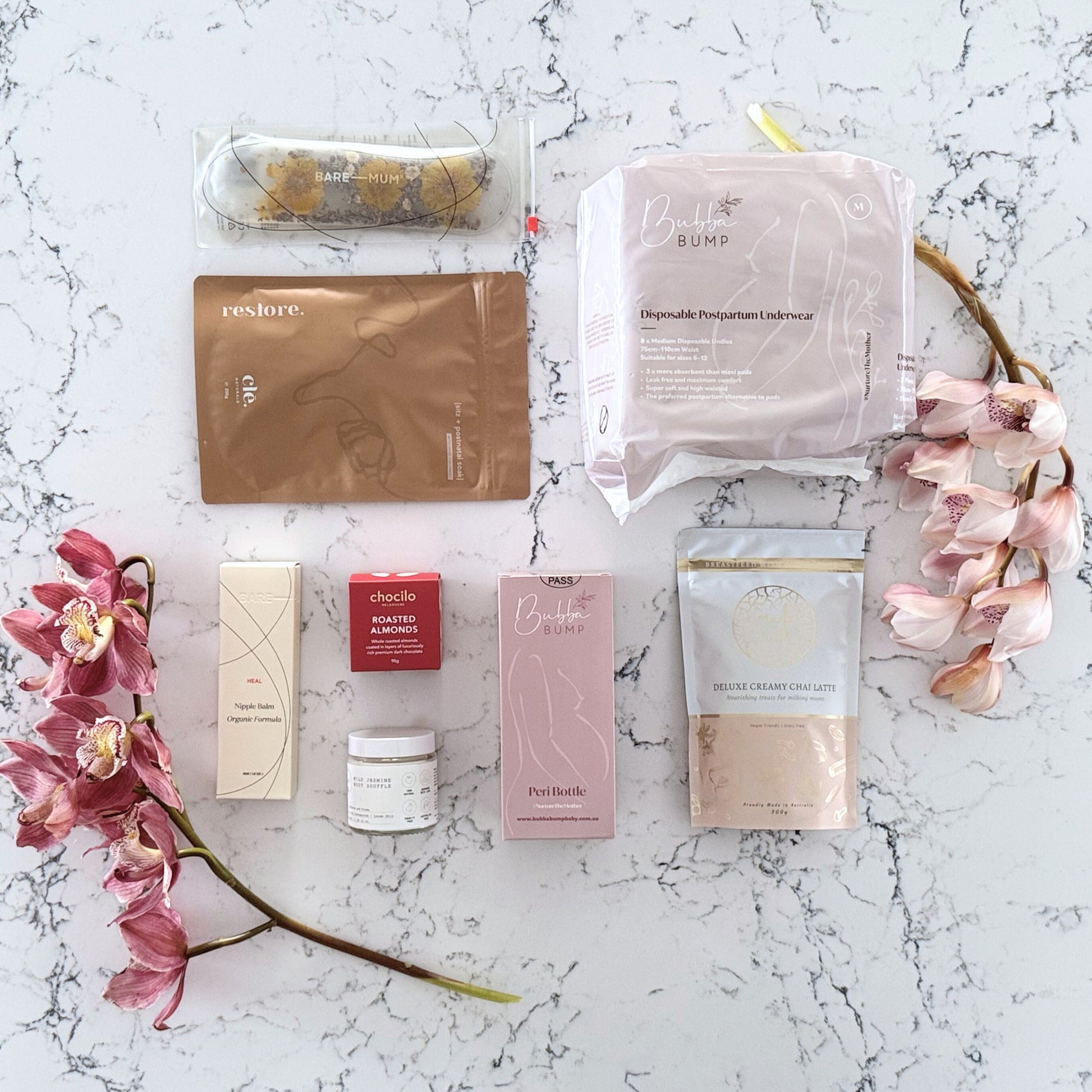 Omoiyari Postpartum Care gift hamper box - Perineal bottle, Sitz bath soak, postpartum underwear, Perineal insert, Creamy Chai latter, Roasted almond chocolate, nipple balm with pink orchid flowers on a marble surface