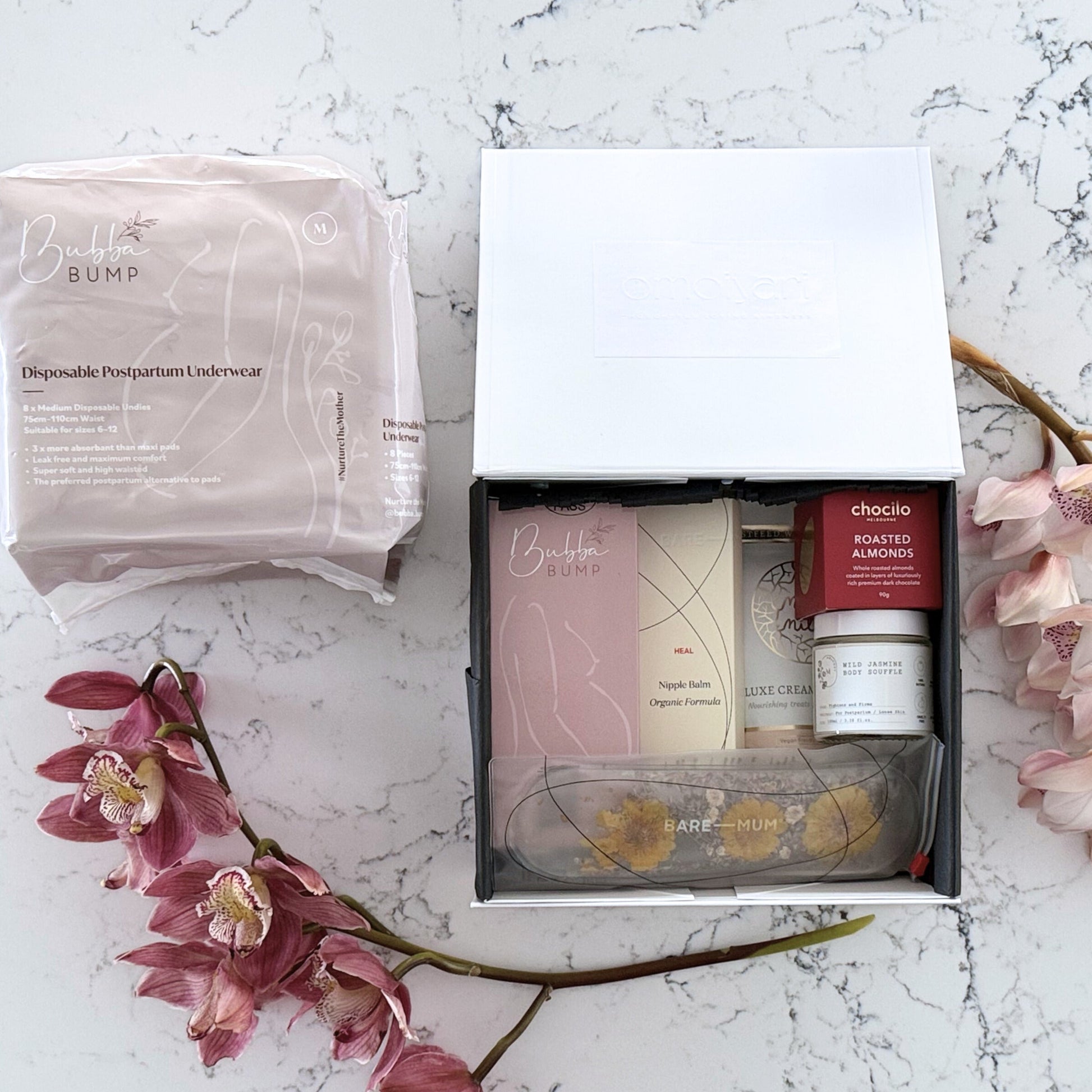 Omoiyari Postpartum Care gift hamper box - Perineal bottle, Sitz bath soak, postpartum underwear, Perineal insert, Creamy Chai latte, Roasted almond chocolate, nipple balm with pink orchid flowers on a marble surface