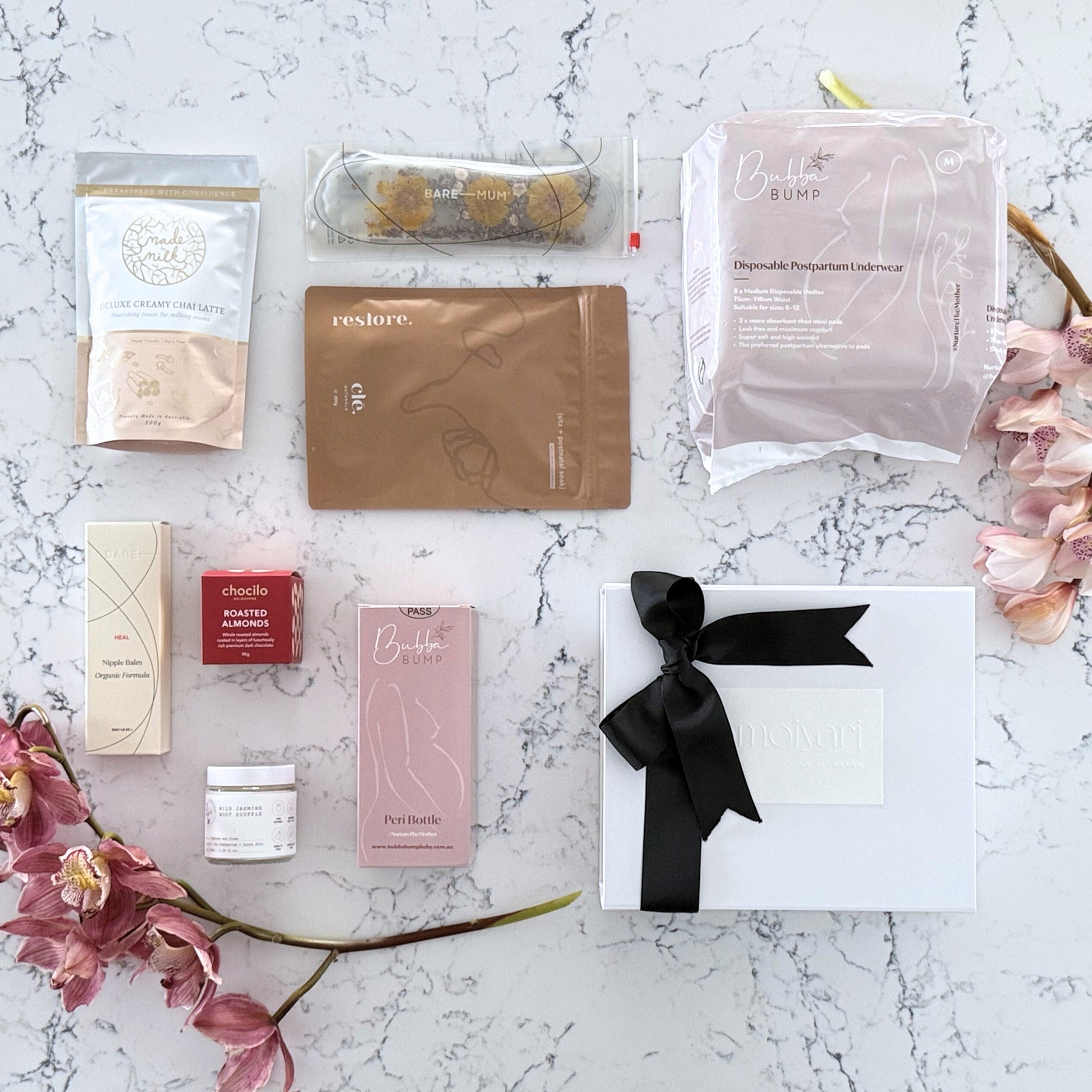 Omoiyari Postpartum Care gift hamper box - Perineal bottle, Sitz bath soak, postpartum underwear, Perineal insert, Creamy Chai latter, Roasted almond chocolate, nipple balm with pink orchid flowers on a marble surface